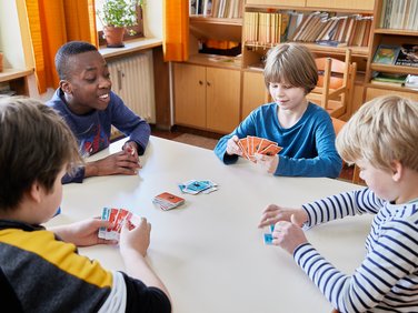 Vier Jungen spielen ein Kartenspiel an einem Tisch in einer Schulbibliothek.