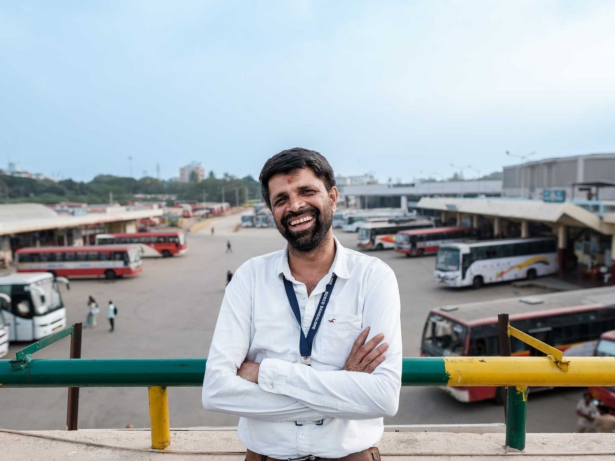 Ein lächelnder Mann in weißem Hemd mit 'BOSCO Bangalore' Lanyard steht mit verschränkten Armen vor einem belebten Busbahnhof.