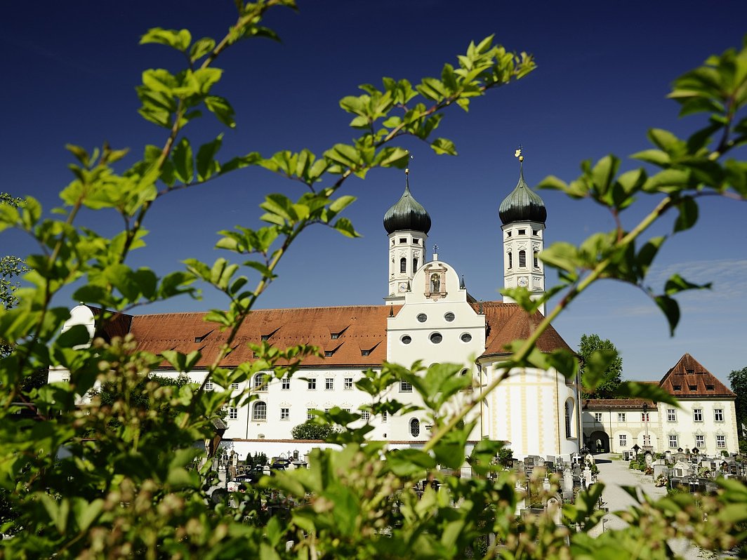 Historische Kirche/Kloster mit zwei Zwiebeltürmen & rotem Dach, umrahmt von unscharfen grünen Blättern. Im Hintergrund ein Friedhof.