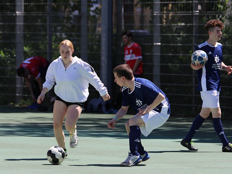 Eine junge Frau in Weiß dribbelt einen Fußball an einem Spieler in Blau vorbei auf einem Außenplatz, andere Spieler sichtbar.