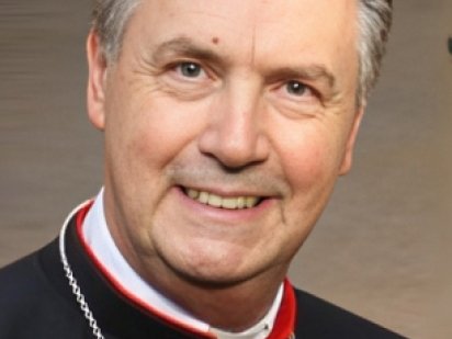Ein lächelnder Kardinal mit rotem Zucchetto und schwarzer Soutane mit rotem Besatz und weißem Priesterkragen.