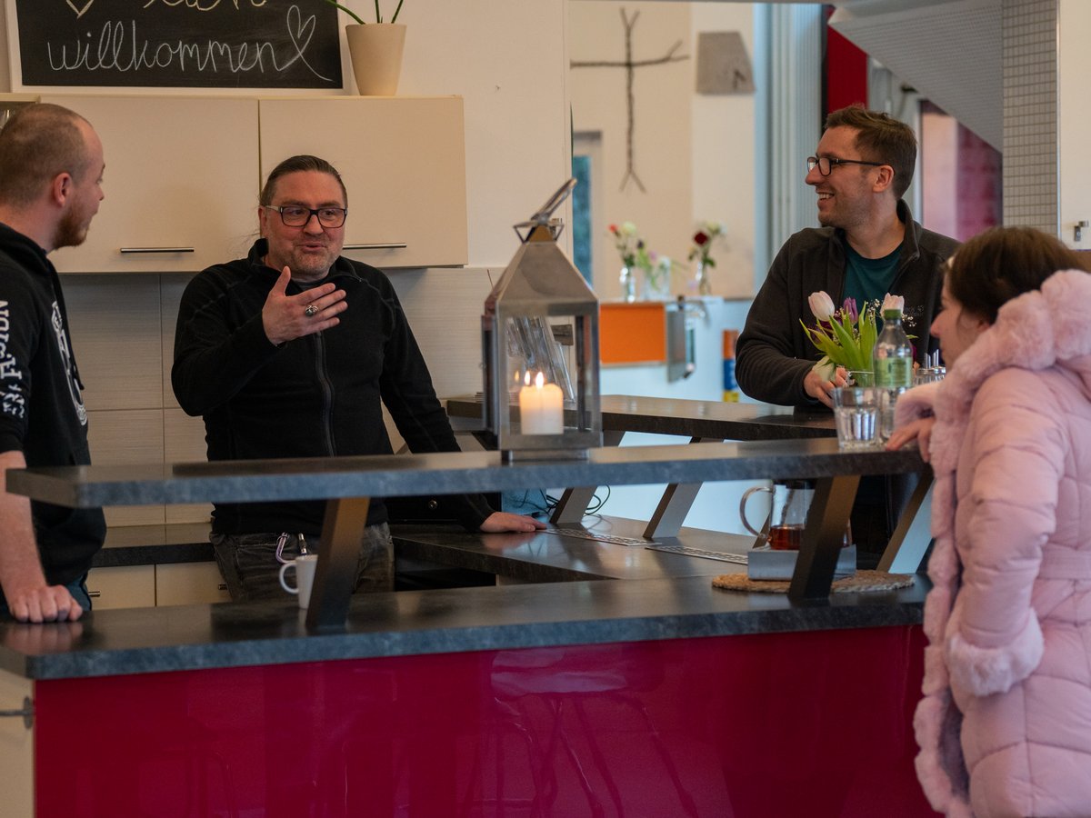 Vier Personen unterhalten sich an einer modernen Küchentheke mit Kerze und Blumen. Im Hintergrund ein Willkommensschild und ein Holzkreuz.