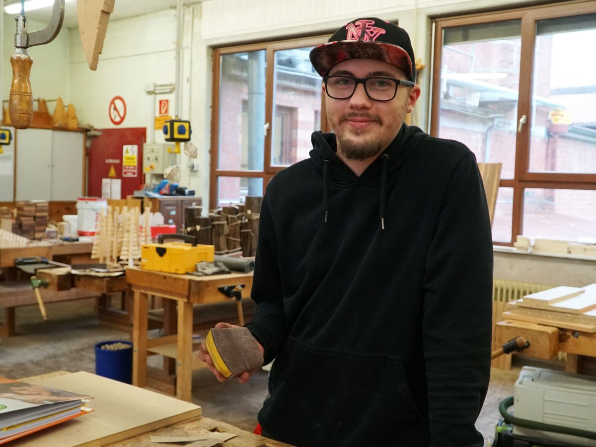 Junger Mann mit Brille und Kappe in einer Holzwerkstatt, hält einen Schleifklotz und lächelt in die Kamera.