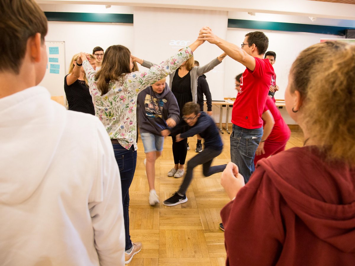 Eine Gruppe von Erwachsenen und Kindern spielt ein aktives Gruppenspiel, bildet einen Kreis mit erhobenen Armen, durch den sich zwei Jungen bewegen.