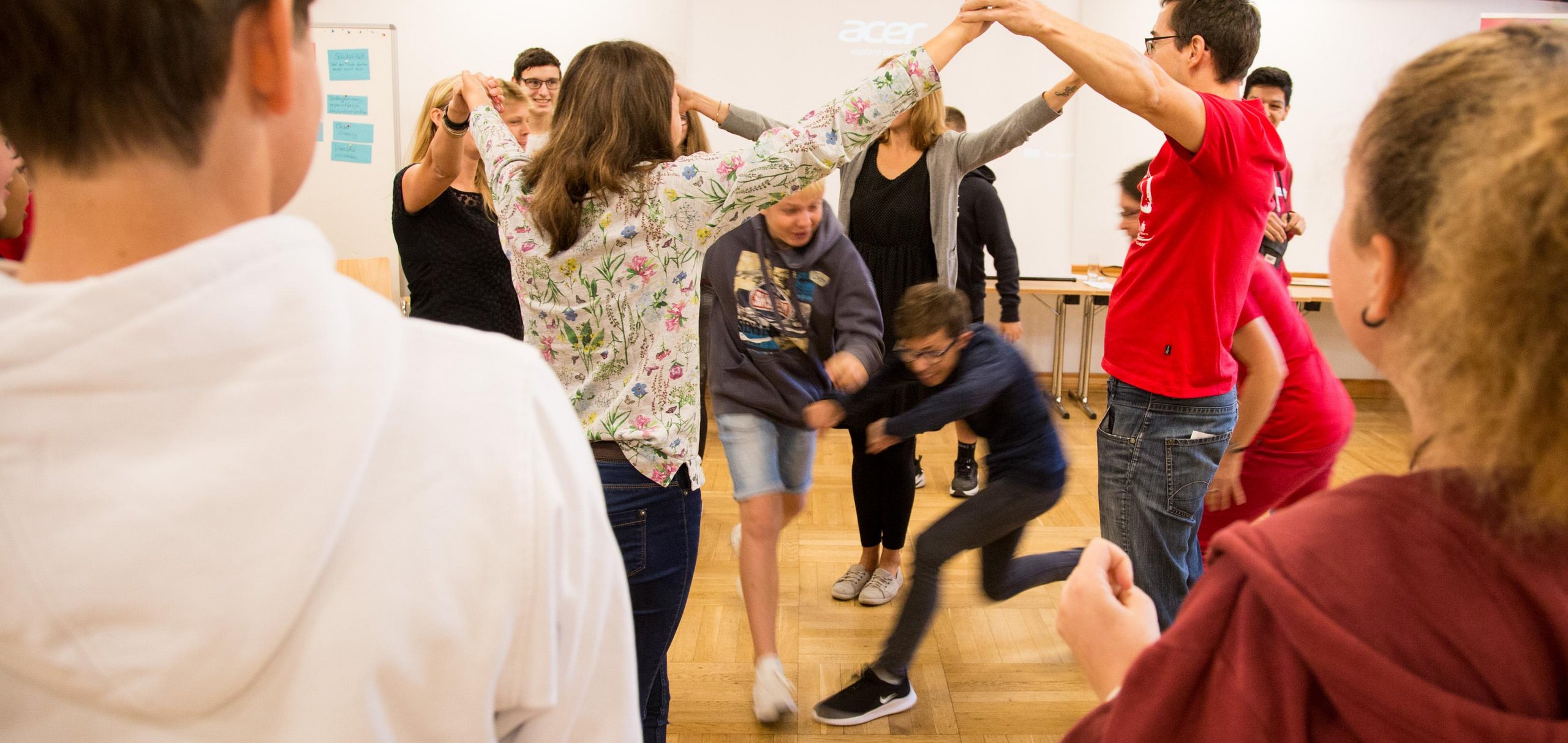 Eine Gruppe von Erwachsenen und Kindern spielt ein aktives Gruppenspiel, bildet einen Kreis mit erhobenen Armen, durch den sich zwei Jungen bewegen.