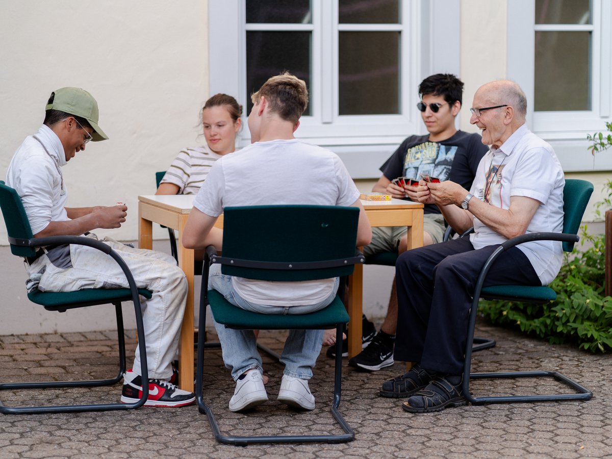 Vier Personen, zwei Generationen, spielen Karten an einem Tisch im Freien. Der ältere Mann lächelt, Karten in der Hand.