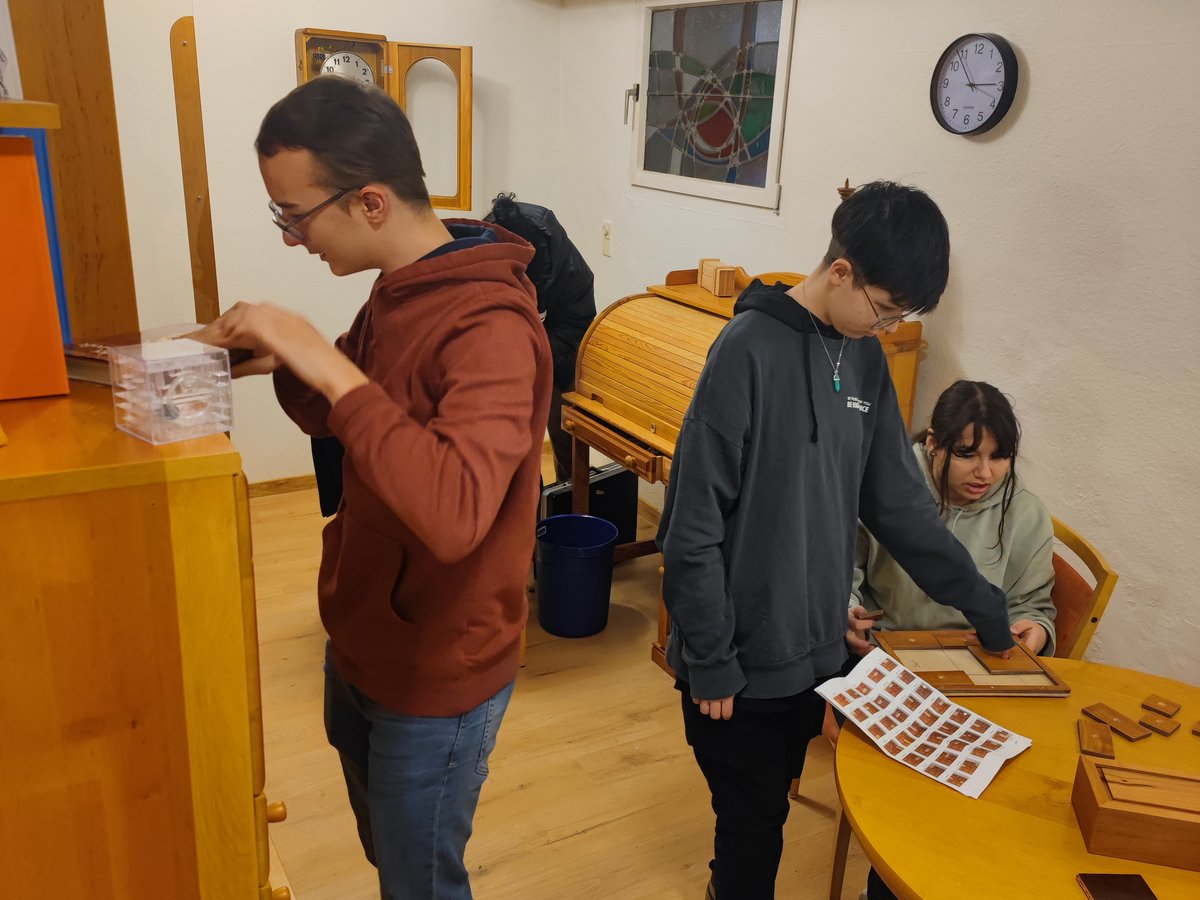 Drei Personen lösen Rätsel in einem Raum: einer mit einem durchsichtigen Würfel, zwei an einem gelben Tisch mit einem Holzschieberätsel.