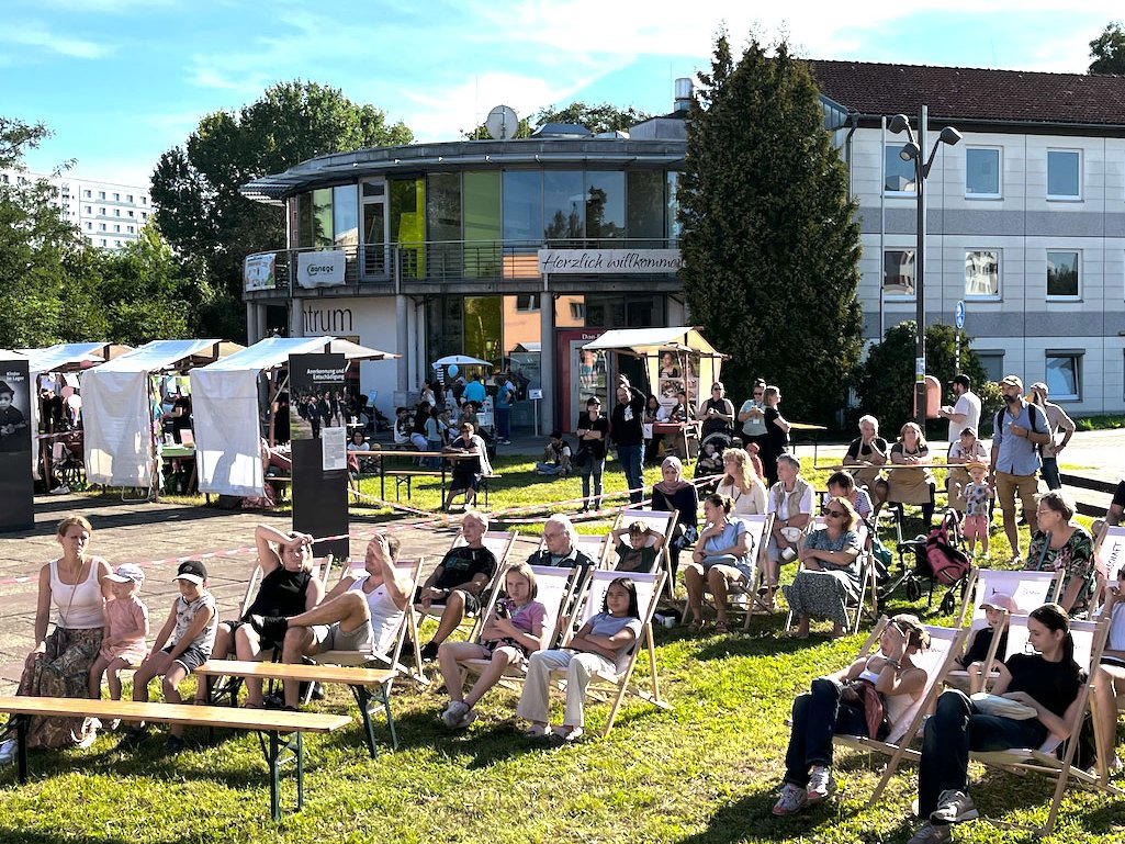 Sonniges Open-Air-Event mit Menschen auf Liegestühlen im Gras, Verkaufsständen und Gebäuden im Hintergrund.