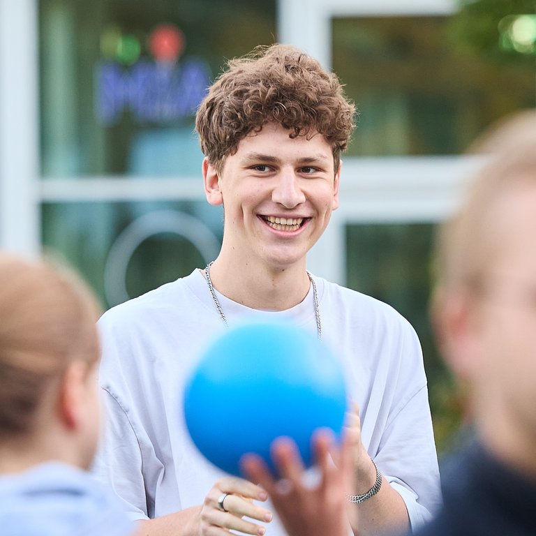 Junger Mann mit lockigem Haar lächelt und hält einen blauen Ball in einer Gruppe von Menschen im Freien.