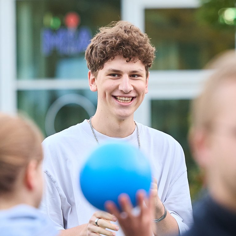 Junger Mann mit lockigem Haar lächelt und hält einen blauen Ball in einer Gruppe von Menschen im Freien.