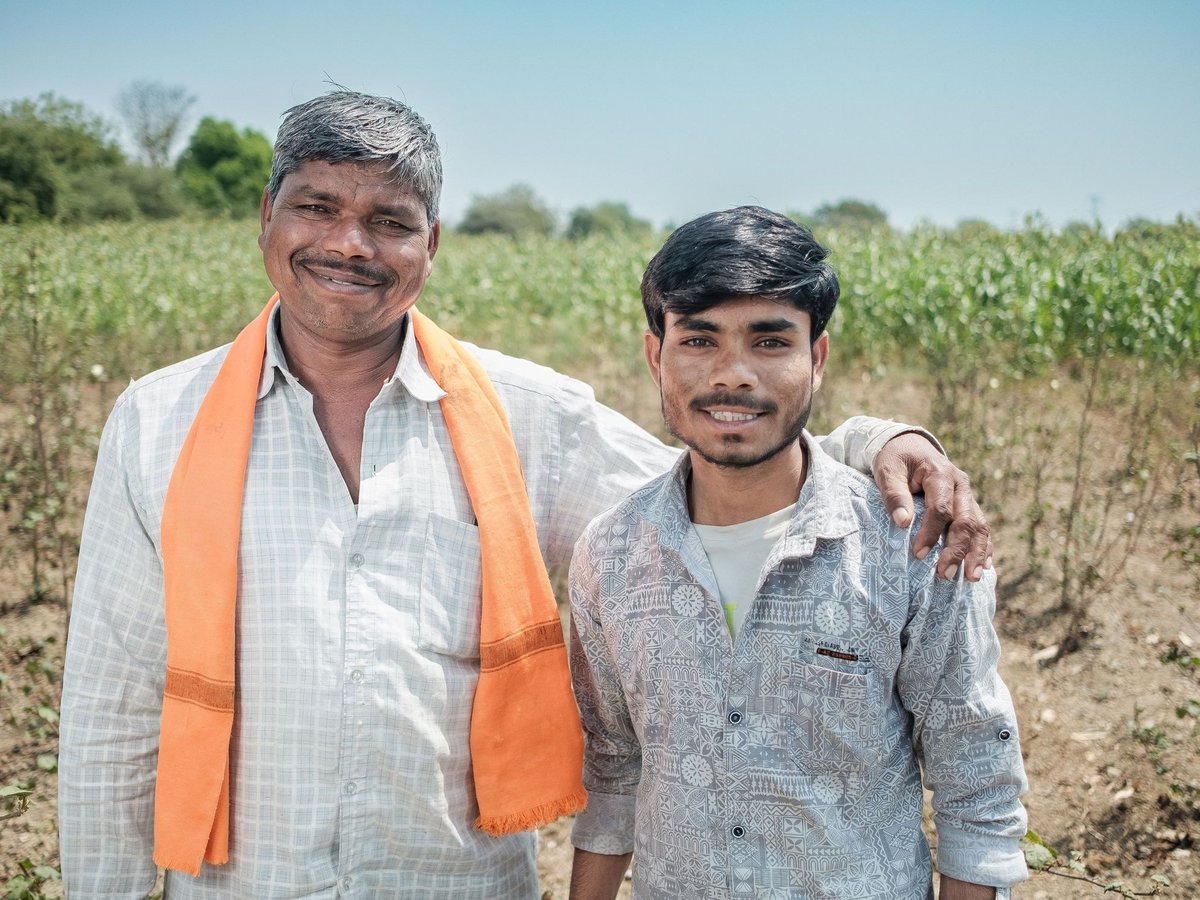 Zwei lächelnde Männer, ein älterer mit dem Arm um einen jüngeren, stehen auf einem grünen Feld unter blauem Himmel.