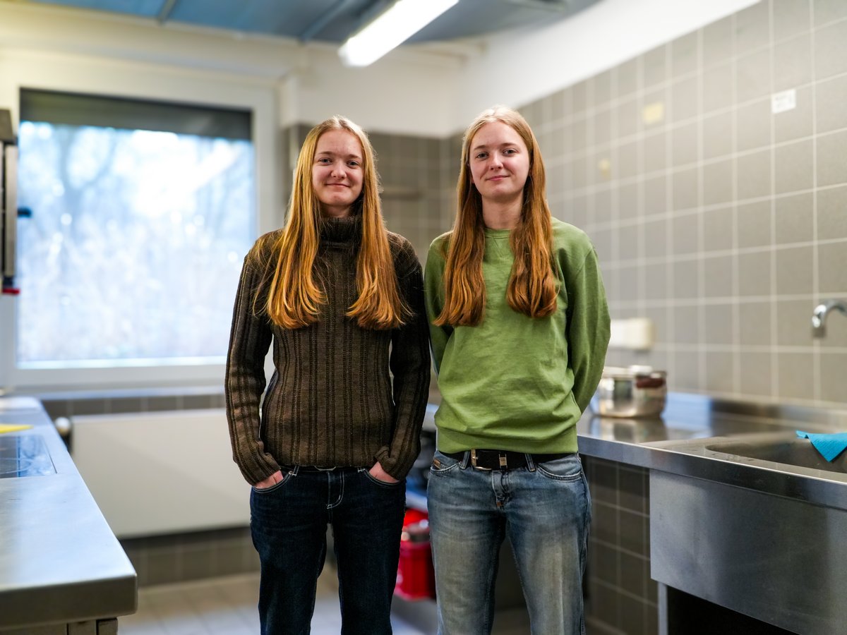 Zwei sehr ähnliche junge Frauen mit langen blonden Haaren lächeln in einer modernen Küche.
