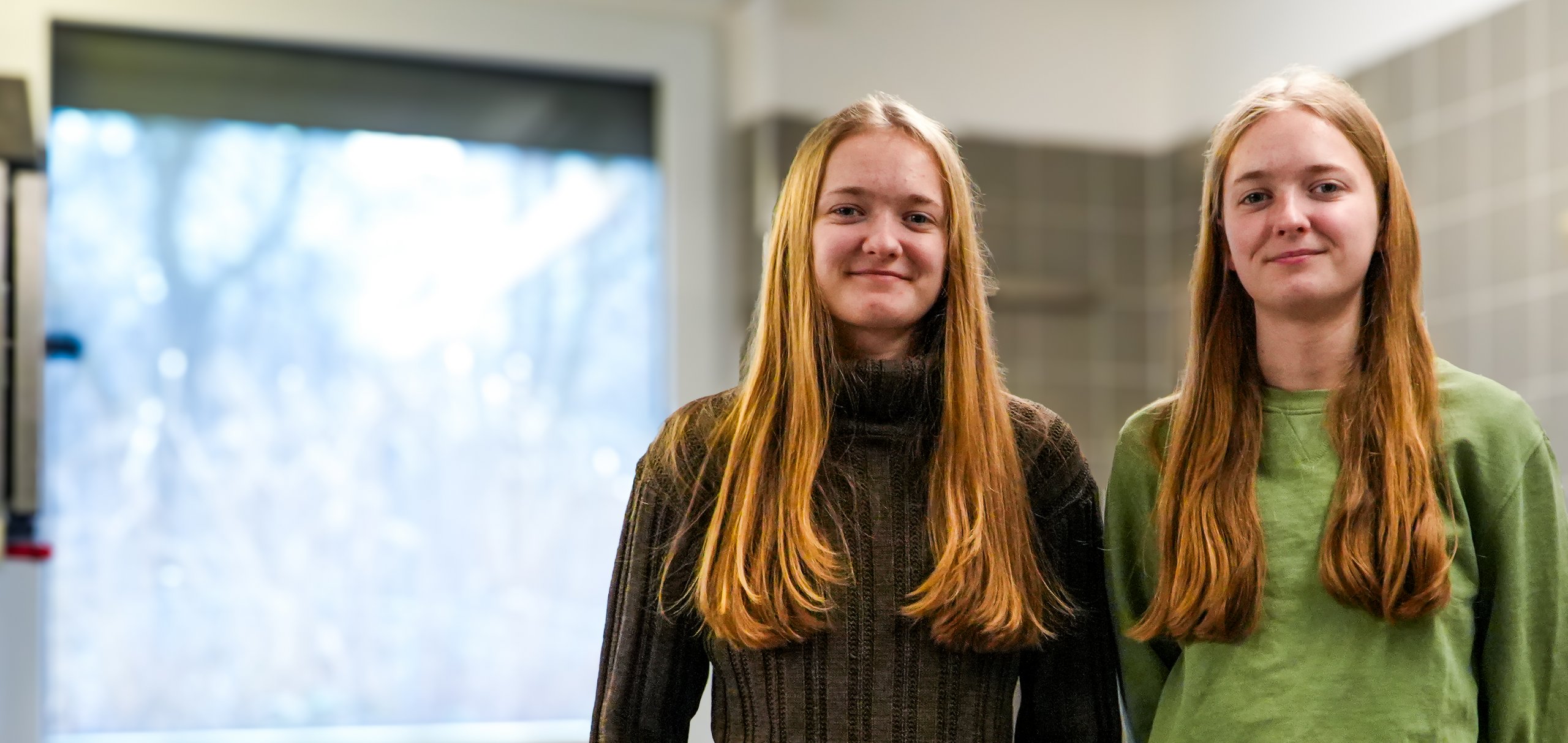 Zwei sehr ähnliche junge Frauen mit langen blonden Haaren lächeln in einer modernen Küche.