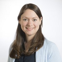 Lächelnde Frau mit langen braunen Haaren, blauen Augen und einem hellblauen Cardigan, blickt direkt in die Kamera.