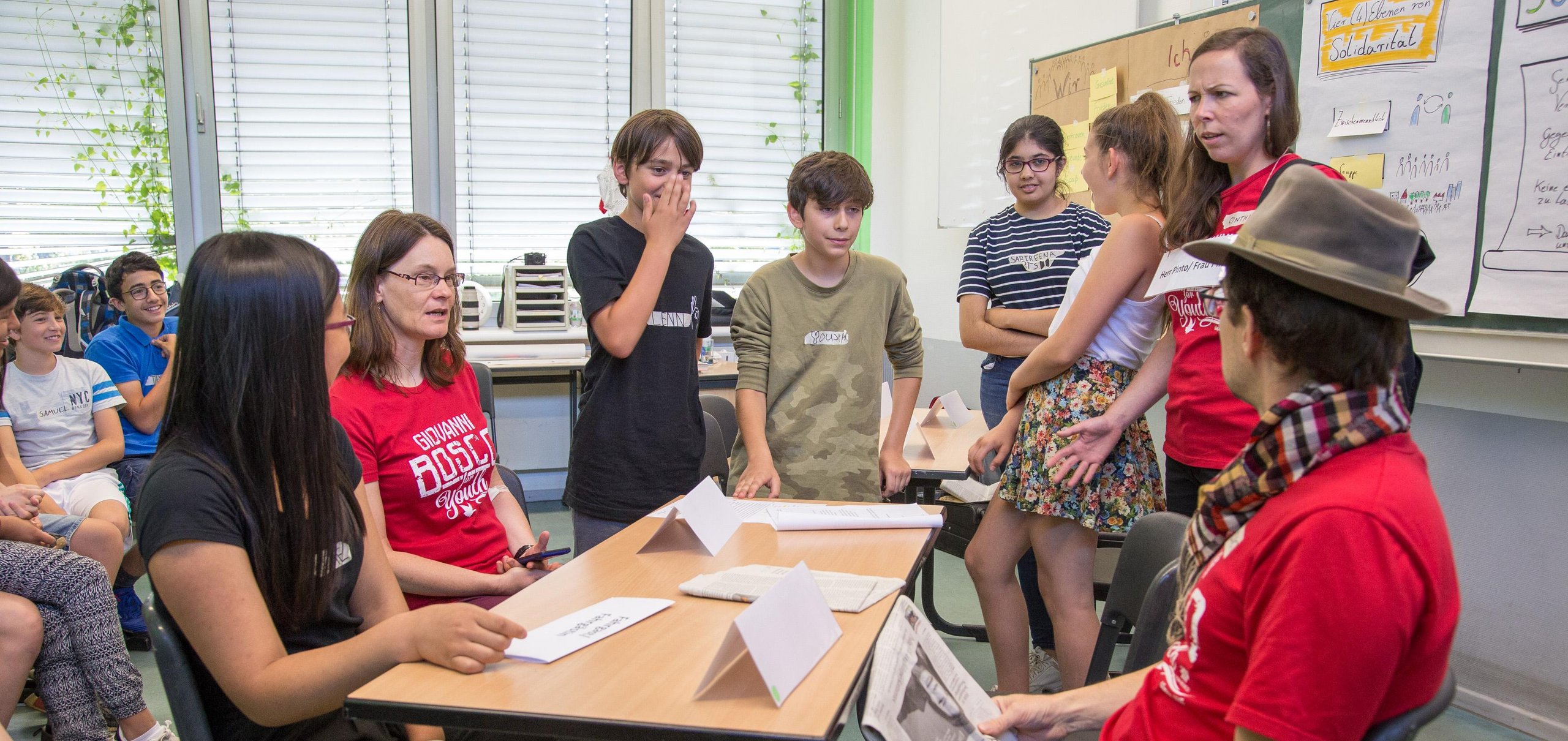Jugendliche und Erwachsene sitzen und stehen in einem Klassenzimmer um einen Tisch, diskutieren, mit Flipcharts im Hintergrund.