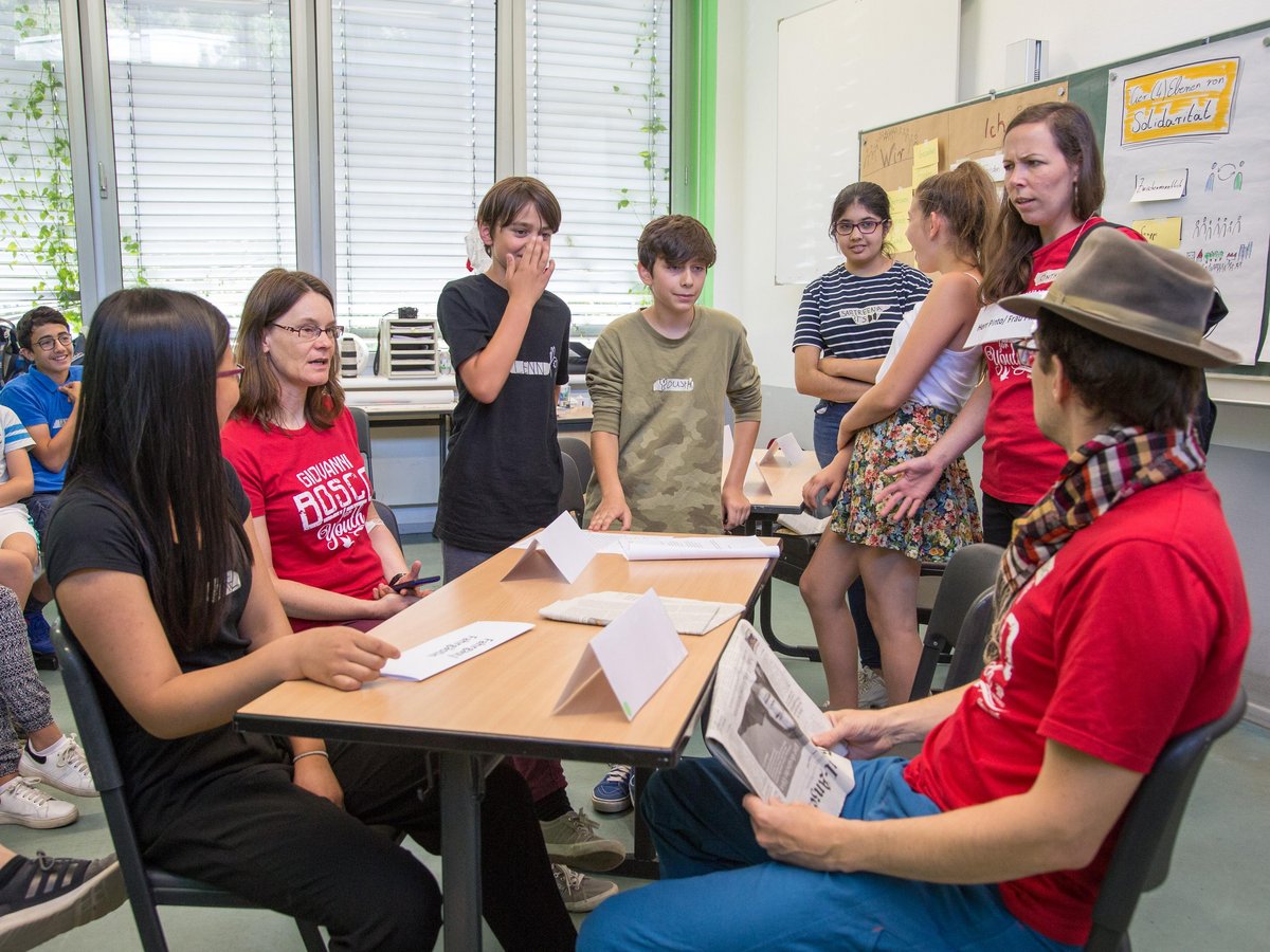 Jugendliche und Erwachsene sitzen und stehen in einem Klassenzimmer um einen Tisch, diskutieren, mit Flipcharts im Hintergrund.