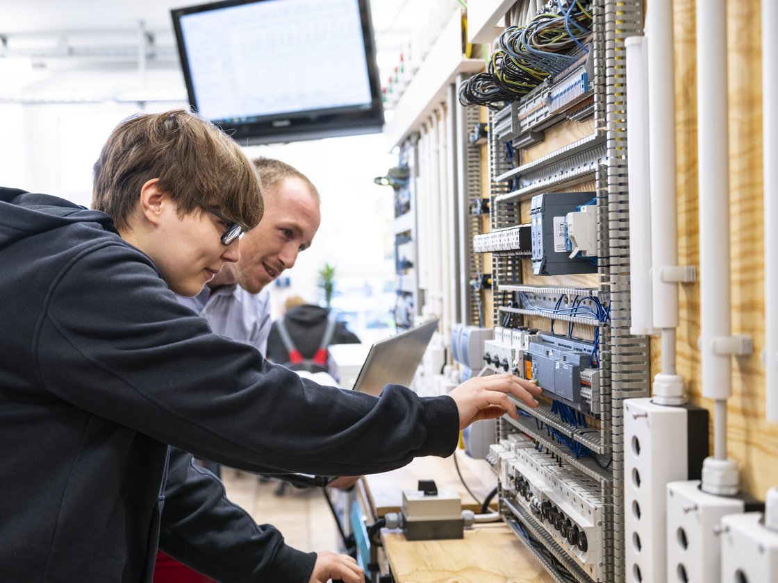 Zwei Männer, einer jung, einer älter, arbeiten an einer elektrischen Schalttafel. Der jüngere zeigt auf ein Bauteil.