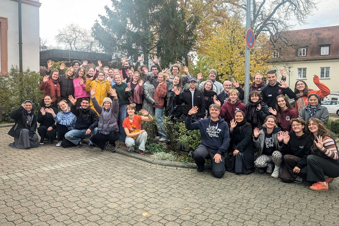 Große Gruppe lachender, winkender Menschen verschiedenen Alters im Freien vor Gebäuden und bunten Herbstbäumen.