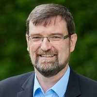 Lächelnder Mann mit grau meliertem Haar, Bart und Brille, in hellblauem Hemd und dunklem Sakko, vor grünem Hintergrund.