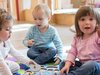 Drei Kleinkinder spielen auf einem bunten Teppich mit Puzzles und Spielzeug; ein Mädchen lächelt in die Kamera und hält einen Stempel.