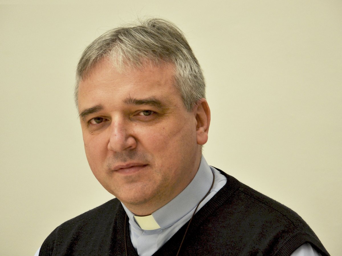 Ein mittelalter Mann mit grauem Haar, Priesterkragen und dunklem Pullover blickt direkt in die Kamera.