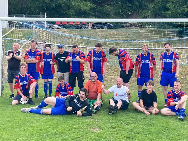 Ein diverses Fußballteam, hauptsächlich Männer in blau-roten Trikots, posiert fröhlich auf einem Rasenplatz vor einem Tor.