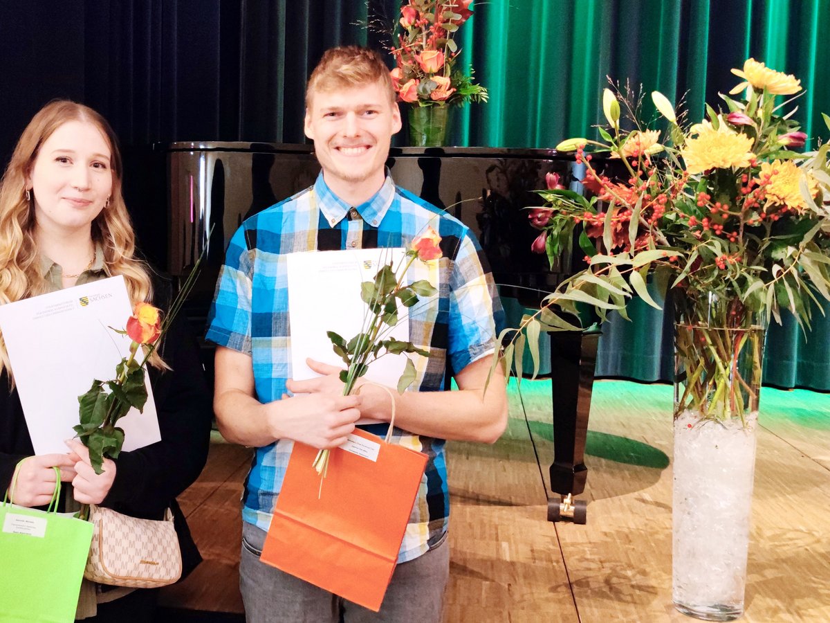 Zwei lächelnde junge Erwachsene halten Urkunden und Rosen auf einer Bühne mit Flügel und grünen Vorhängen im Hintergrund.