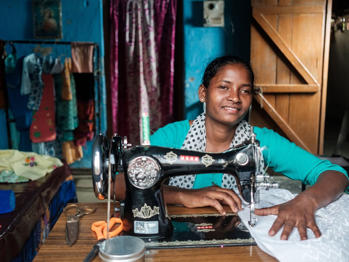 Lachende Frau näht an einer traditionellen Nähmaschine. Ihr Atelier ist mit bunten Stoffen geschmückt.