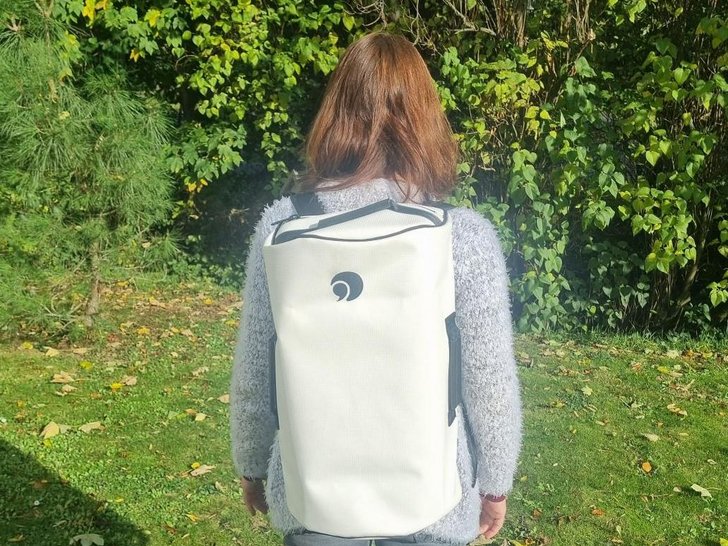 Person von hinten mit einem hellgrauen flauschigen Pullover und einem cremefarbenen Rucksack mit schwarzem Spiral-Logo im Garten.