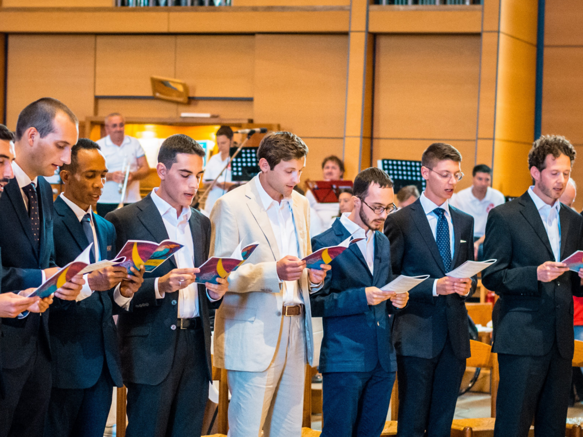 Junge Männer in Anzügen singen aus Liederbüchern in einem feierlichen Rahmen, vermutlich einer Kirche.