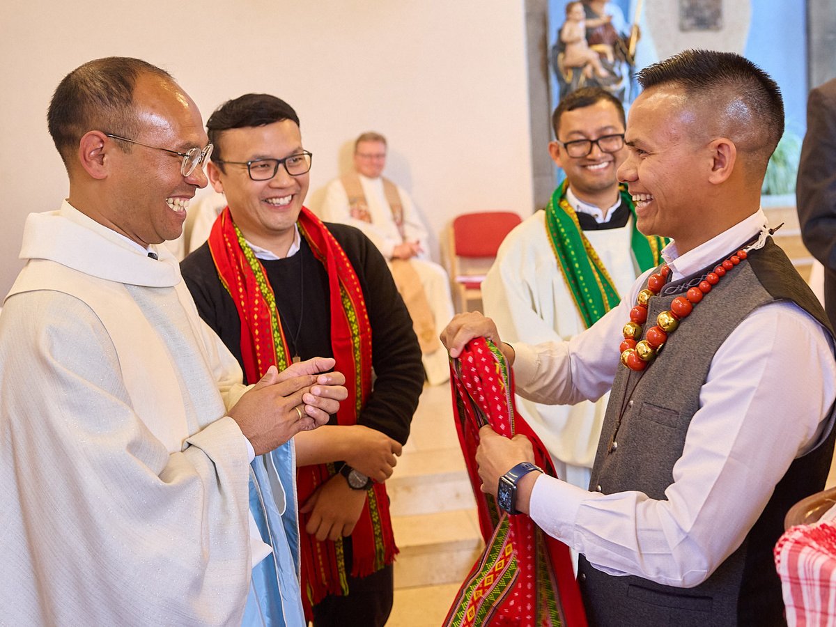 Drei Männer in einer Kirche, einer im Priestergewand, einer mit rotem Schal und einer mit roter Perlenkette, alle lächeln fröhlich.