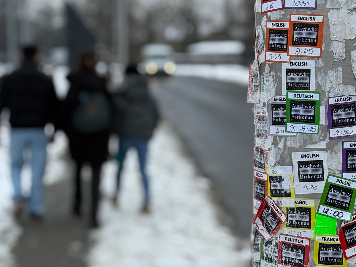 Ein mit Flyern für Auschwitz-Birkenau-Touren in vielen Sprachen bedeckter Pfosten. Im Hintergrund gehen Personen auf einem schneebedeckten Weg.