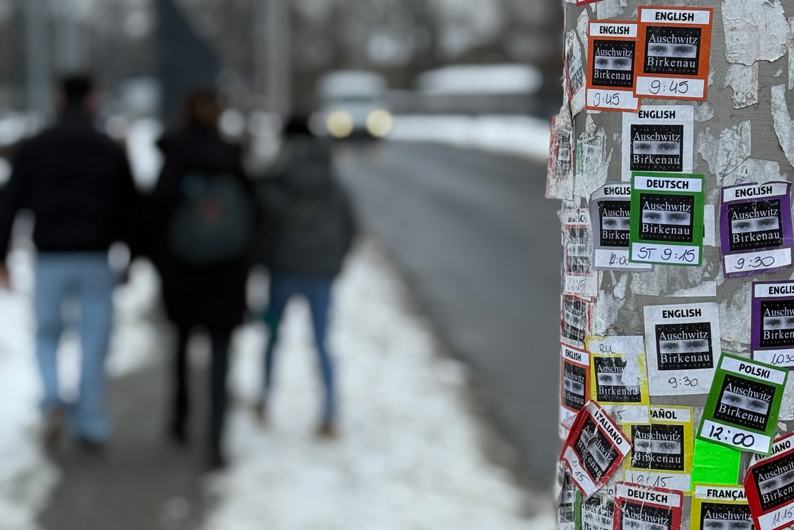 Ein mit Flyern für Auschwitz-Birkenau-Touren in vielen Sprachen bedeckter Pfosten. Im Hintergrund gehen Personen auf einem schneebedeckten Weg.