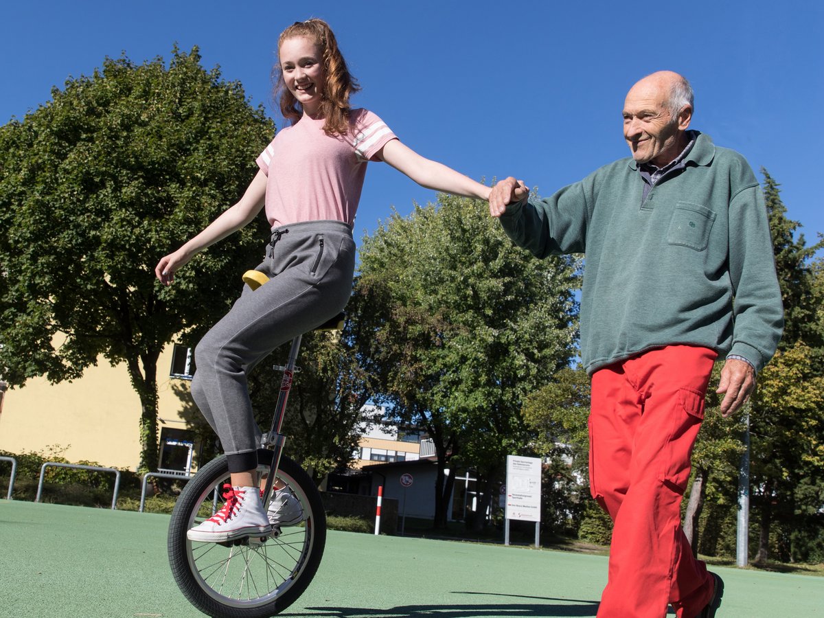 Eine junge Frau auf einem Einrad hält Händchen mit einem älteren Mann, der ihr lächelnd auf einem grünen Platz hilft.