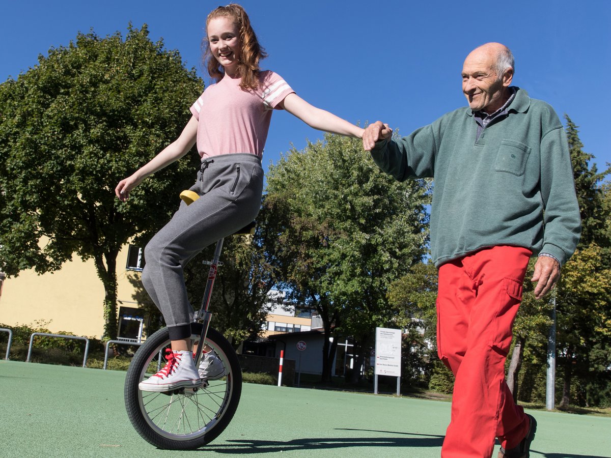 Eine junge Frau auf einem Einrad hält Händchen mit einem älteren Mann, der ihr lächelnd auf einem grünen Platz hilft.