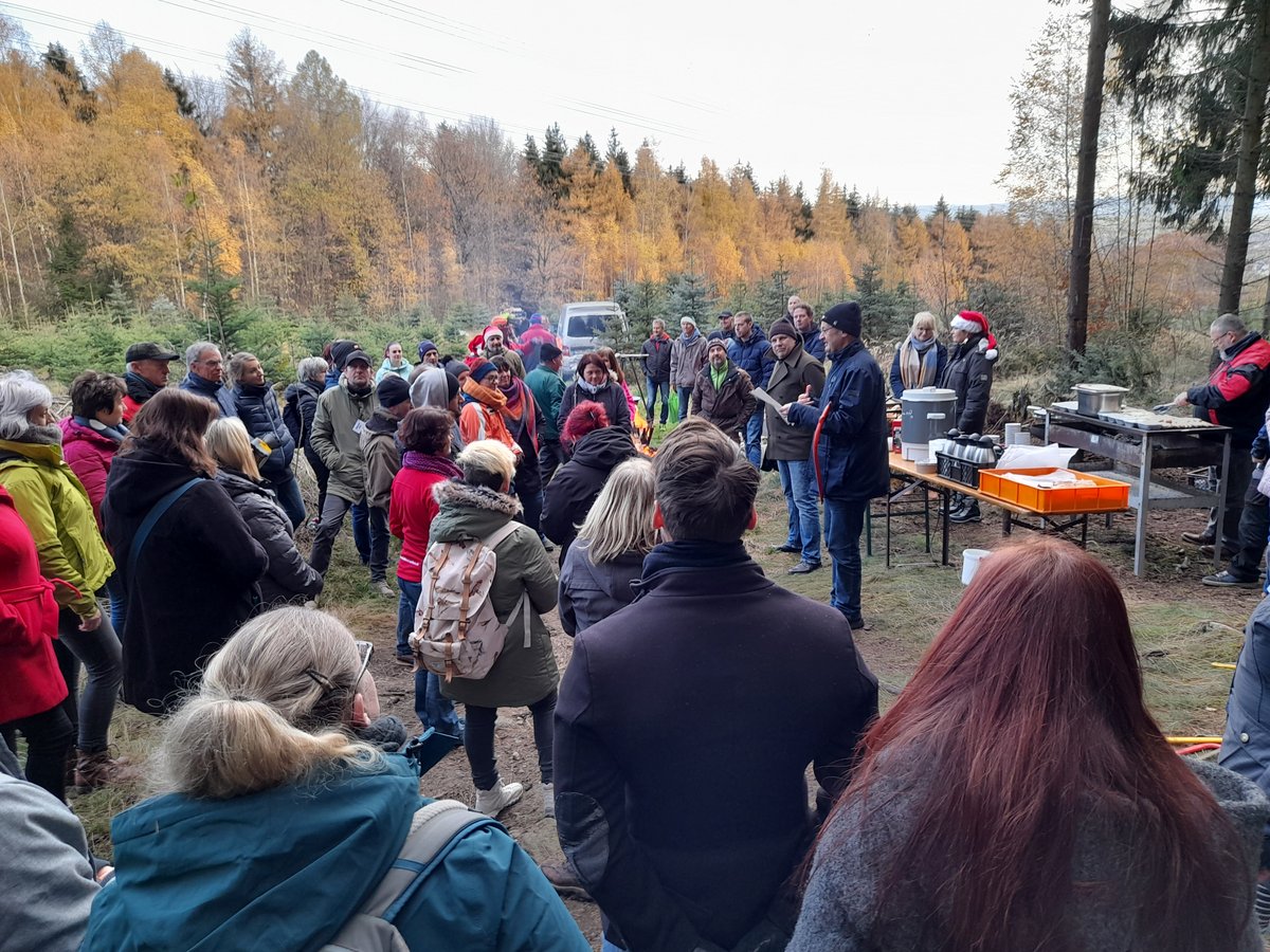 Menschen treffen sich im Herbstwald um ein Lagerfeuer, hören einem Redner zu und genießen Essen vom Grill.