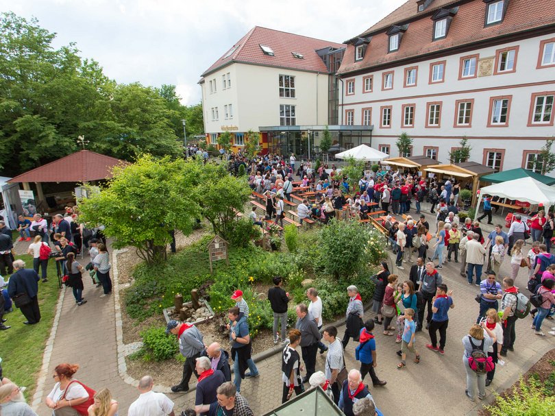 Viele Menschen bei einer lebhaften Outdoor-Veranstaltung in einem grünen Gartenhof mit Gebäuden, Bänken und Ständen.