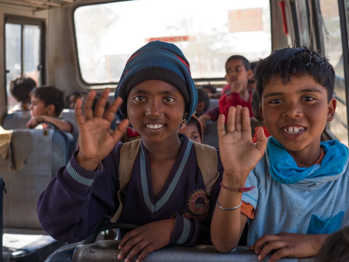 Zwei lächelnde Kinder, eines mit Mütze und eines mit Kapuzenpullover, winken aus einem belebten Bus.