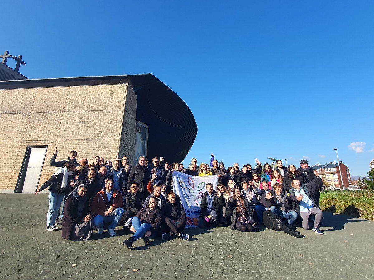 Eine große, vielfältige Gruppe lächelnder Menschen posiert im Freien mit einem 'Salesian Youth'-Banner vor einer modernen Kirche.