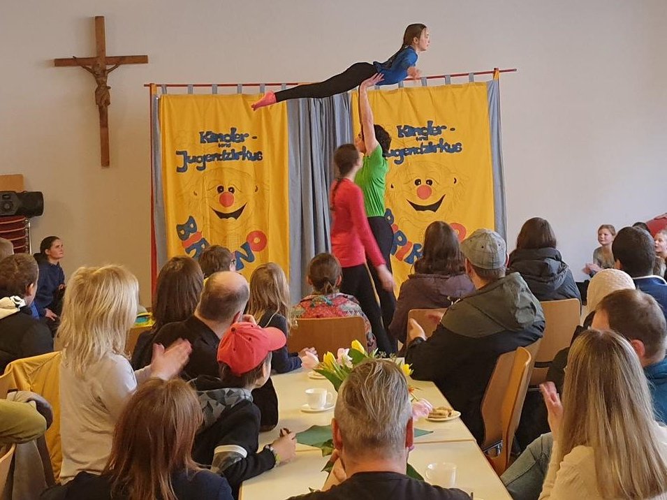 Zwei Personen des Kinder- und Jugendzirkus führen Akrobatik vor einem sitzenden, klatschenden Publikum in einem Saal auf.