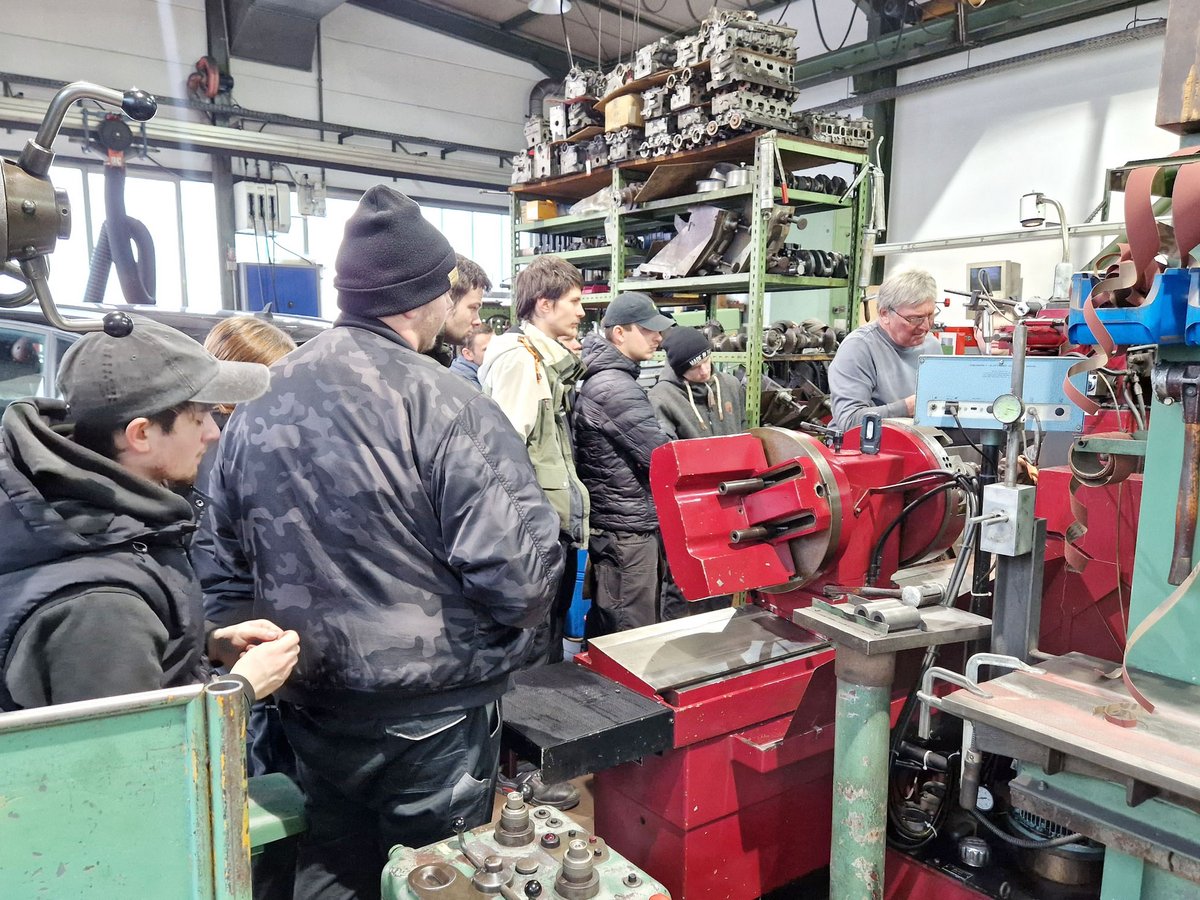 Gruppe von Männern beobachtet Maschinen in einer Werkstatt, älterer Mann demonstriert, Regale mit Motorteilen im Hintergrund.