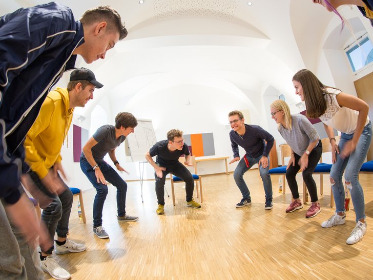 Junge Erwachsene in einem Kreis, kauernd und nach innen blickend bei einer lebhaften Teamaktivität.