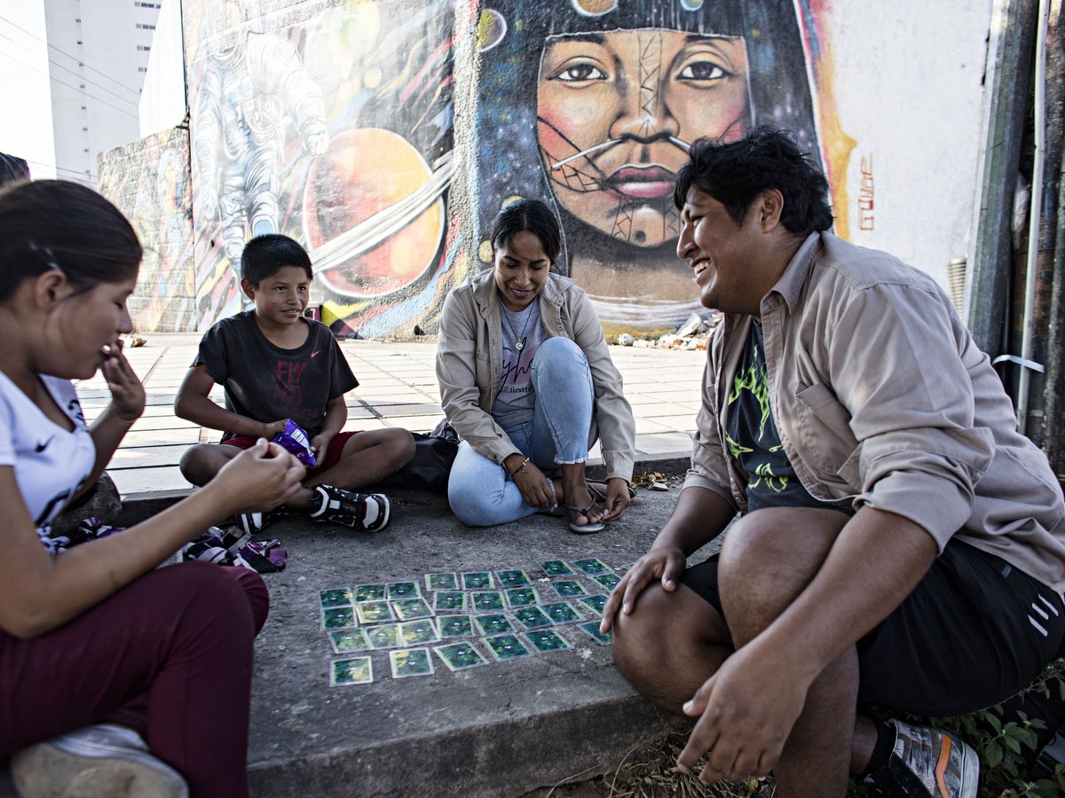 Vier Personen spielen ein Kartenspiel auf dem Boden vor einer bunten Graffitiwand mit einem indigenen Gesicht, Astronauten und Planeten.