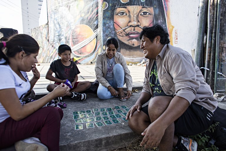 Vier Personen spielen ein Kartenspiel auf dem Boden vor einer bunten Graffitiwand mit einem indigenen Gesicht, Astronauten und Planeten.