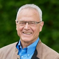 Lächelnder älterer Mann mit weißen Haaren und Brille, blauem Hemd und brauner Jacke, vor verschwommenem grünen Hintergrund.