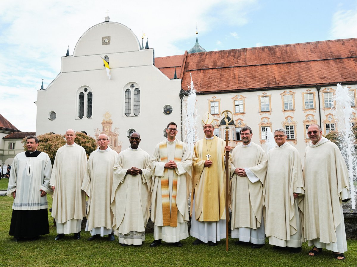 Neun katholische Kleriker, darunter ein Bischof, stehen vor einem imposanten Klostergebäude mit Springbrunnen auf einer Wiese.