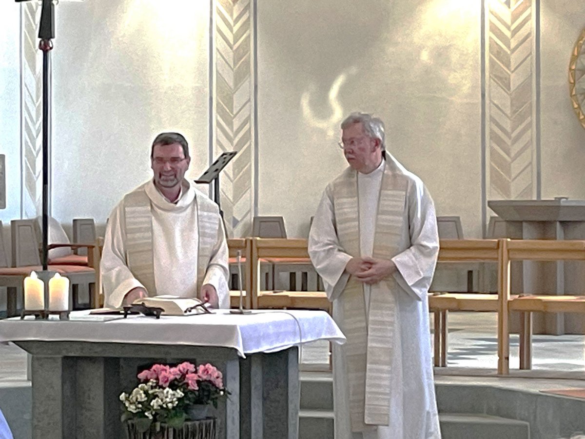 Zwei Priester in weißen Gewändern leiten einen Gottesdienst in einer Kirche. Einer steht lächelnd am Altar mit Buch und Kerzen.