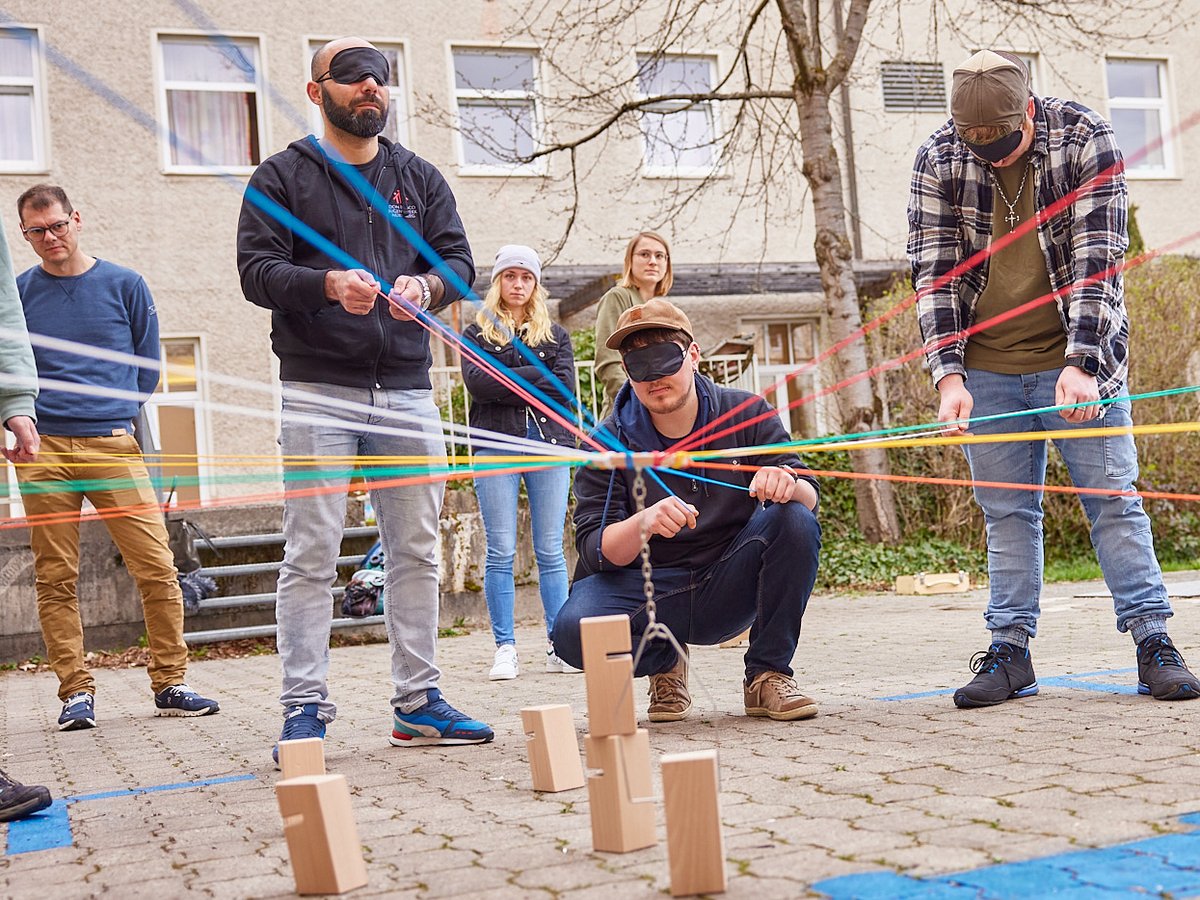 Blinde Menschen versuchen mit bunten Fäden und einem Haken Holzklötze zu stapeln, unterstützt von sehenden Beobachtern.