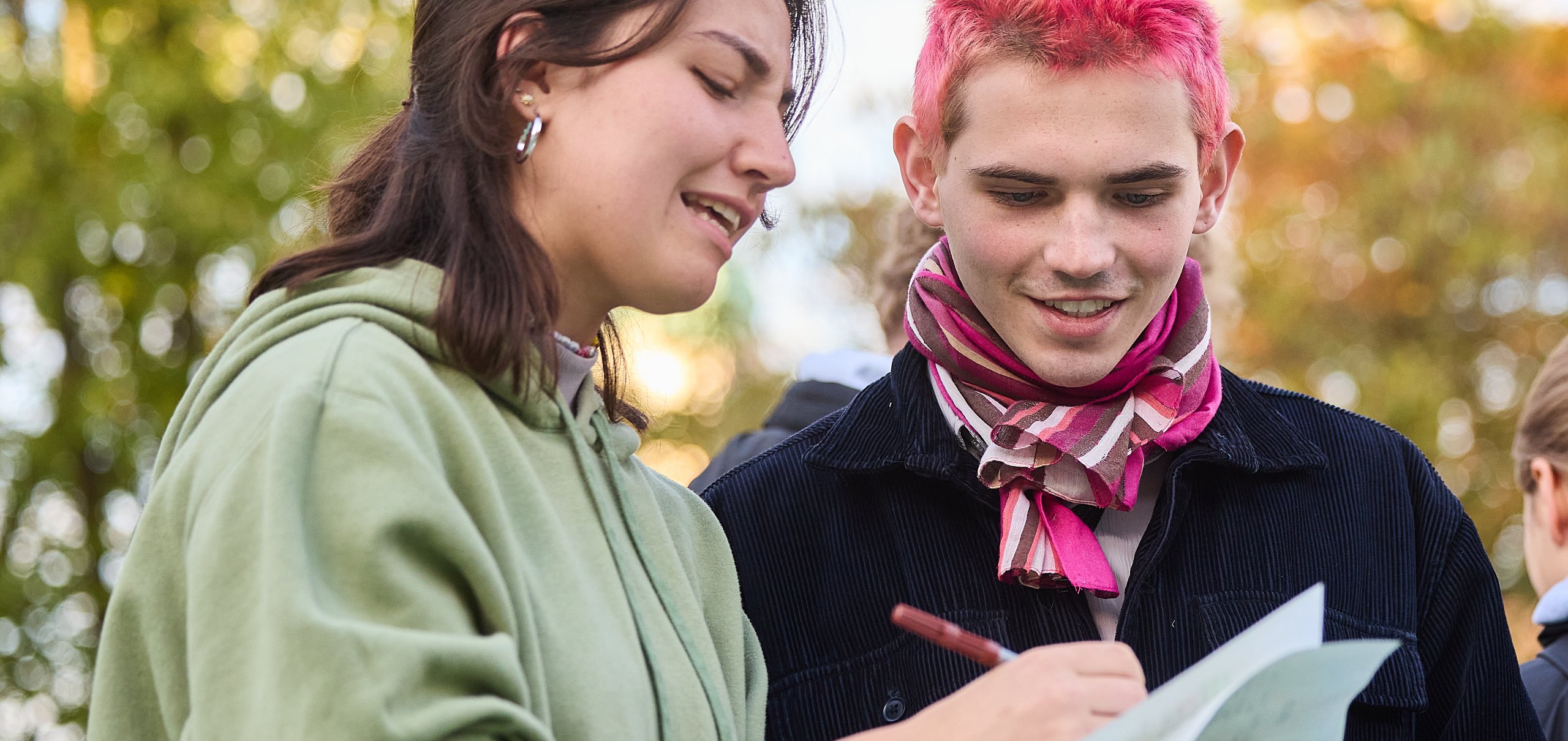 Eine junge Frau und ein junger Mann mit pinken Haaren arbeiten draußen an Papieren, umgeben von Herbstlaub.