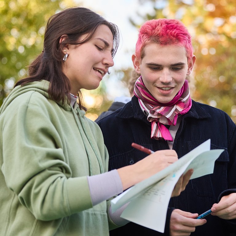 Eine junge Frau und ein junger Mann mit pinken Haaren arbeiten draußen an Papieren, umgeben von Herbstlaub.